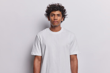 Waist up shot of serious curly haired Hindu man looks directly at camera keeps arms down has confident expression dressed in casual t shirt isolated on white background. Human face expressions concept