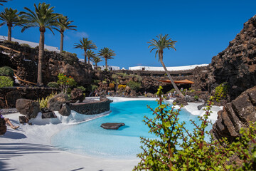 Pool of blue waters and palemeeras, outside the cave of Los Jameos del Agua. Light at the end of the cave. Sky with big white clouds. Lanzarote, Canary Islands, Spain.