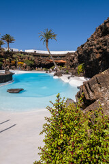 Pool of blue waters and palemeeras, outside the cave of Los Jameos del Agua. Light at the end of the cave. Sky with big white clouds. Lanzarote, Canary Islands, Spain.