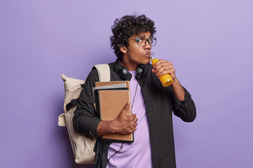 Worried curly haired Hindu student drinks fresh orange juice and carries spiral notepads wears...