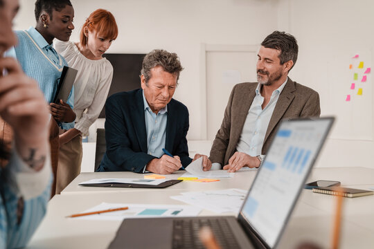 Senior executive signing a contract with a multigenerational team observing, in a collaborative office environment.