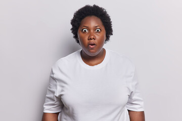 Indoor shot of stunned African woman with curly hair stares bugged eyes cannot believe in shocking news feels amazed dressed in casual jumper isolated over white background. Human reactions concept
