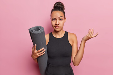 Hesitant doubtful Latin sportswoman has healthy body habits holding rolled fitness mat shrugs shoulders with doubt wears black tracksuit isolated over pink background. Sporty lifestyle concept - obrazy, fototapety, plakaty