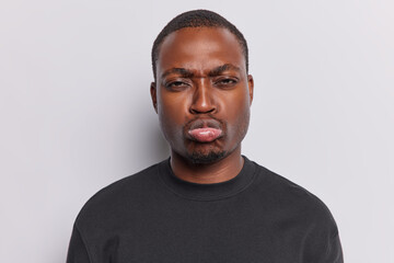 Troubled offended dark skinned man has sulking expression purses lips feels unhappy has some problems in life being hurt by bad words dressed in casual basic black t shirt isolated on white background