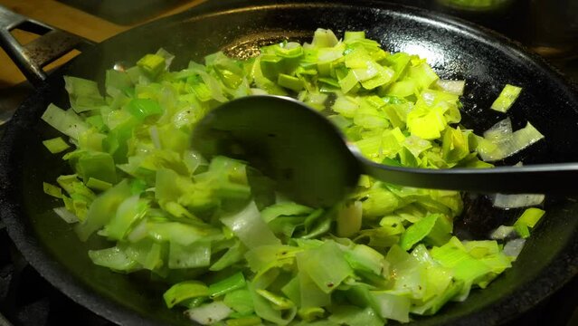Closeup of a large spoon stirring steaming hot, chopped green leeks, frying in a pan.