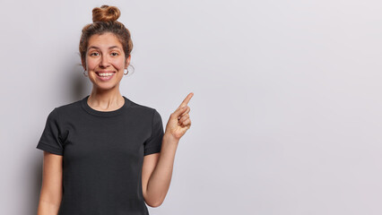 Fototapeta premium Horizontal shot of pretty young European woman with hair bun smiles pleasantly dressed in casual black tshirt points index finger on empty space isolated over white background. Look at this.