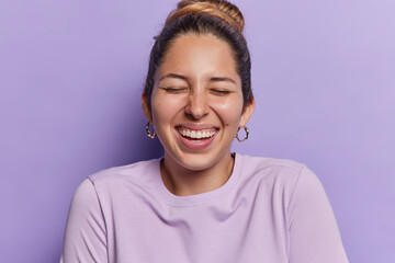Happy sincere woman with hair gathered in bun keeps eyes closed smiles toothily expresses positive emotions and feelings dressed in casual pullover isolated over purple background. Happiness concept