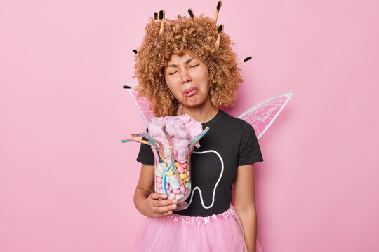 Studio shot of upset curly haired European woman holds glass full of cotton candy and sweets dressed in costume of tooth fairy has toothbrush stuck in curly hair isolated over pink background