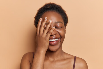 Horizontal shot of happy dark skinned African woman with curly short hair makes face palm keeps...