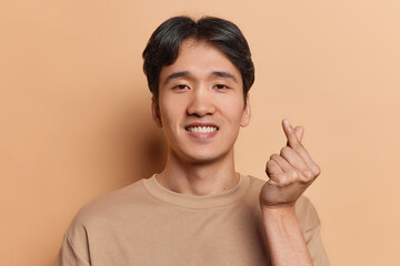 Handsome dark haired Chinese man smiles pleasantly shows mini heart gesture expresses love dressed in casual t shirt shows korean like sign isolated over brown background. Body language concept