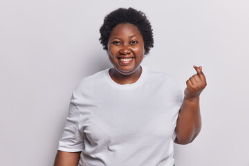 Happy dark skinned overweight African woman with curly hair makes korean like gesture shows mini...