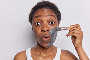 Surprised dark skinned woman puts on clay mask with cosmetic brush for skinn moisturising keeps...
