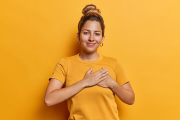 Horizontal shot of pretty young woman presses hands to heart expresses gratitude expressing appreciation and kindness smiles happily dressed in casual t shirt isolated over yellow background - obrazy, fototapety, plakaty