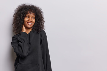 Horizontal shot of pretty curly haired woman keeps hand on neck smiles gently focused aside has dreamy expression wears black sweatshirt being satisfied with something isolated over white background