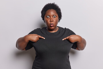Waist up shot of shocked African woman points at blank space of casual basic tshirt shows mock up...
