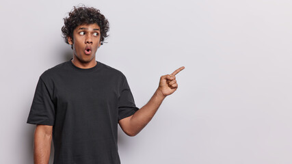 People and emotions. Indoor photo of young surprised Hindu male standing on left isolated on white background pointing and looking at blank space for your advertisement wearing black t shirt - Powered by Adobe