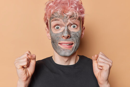 Photo of surprised pink haired man applies facial clay mask for skin moisturising clenches fists awaits for something happened dressed in casual black t shirt isolated over brown background.