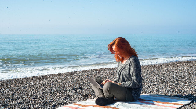 A young red-haired woman works on a laptop remotely on the beach by the sea. High quality photo