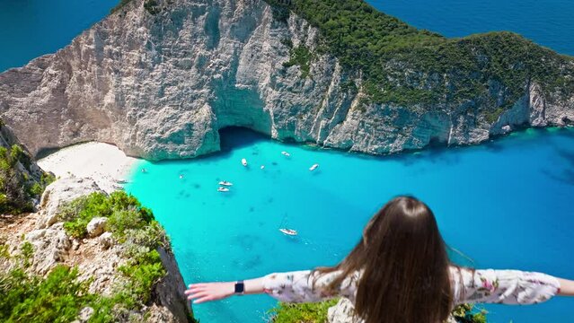 Aerial view of a beautiful woman looking at Navagio Pebble Beach with a shipwreck and turquoise water. Female tourists enjoying summer vacation at the cove reached by boats in Zakynthos, Greece.