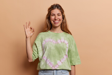 Pretty young woman shows okay gesture says everything alright has situation under control guarantees all is good dressed in casual clothing isolated over brown background. Body language concept