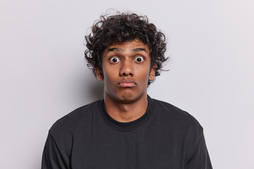Human reactions concept. Stunned curly haired Hindu man purses lips and stares with bugged eyes at...