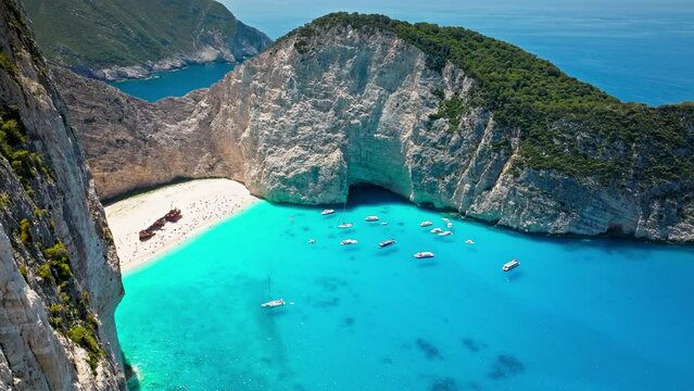 Navagio Pebble Beach between towering cliffs with a shipwreck and turquoise water. Aerial view of tourists enjoying summer vacation at the cove reached by boats, Navagio Beach with crystal blue water.
