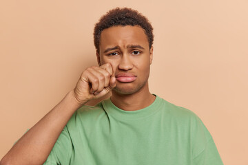 People negative emotions concept. Studio portrait of upset African american man standing in centre isolated on beige background looking as if crying and wiping tears holding fist under left eye