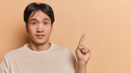Young handsome brunet young Japanese man has wondered expression points index finger on blank space for your promotion dressed in casual t shirt isolated over brown background. Wow look at this