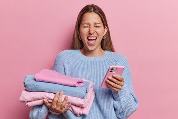 Photo of emotional long haired woman holding neatly folded pile of clothes and smartphone wears knitted jumper isolated over pink background surfs resale fashion sites. Housekeeping concept.