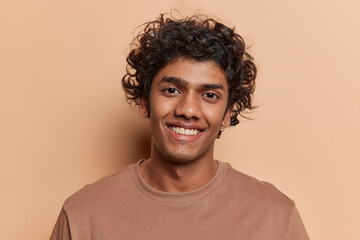 Portrait of merry positive curly haired Hindu man smiles pleasantly being in good mood dressed in casual t shirt looks directly at camera isolated over brown background. People and emotions concept