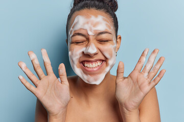 Studio shot of cheerful young woman uses cleansing foam keeps eyes closed smiles toothily keeps...