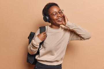 Happy joyful African girl returns home from school enjoys listening muic via headphones catches...