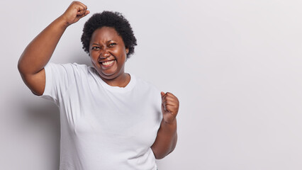 Photo of overjoyed dark skinned plump woman makes winner gesture raises arms and clenches fists...