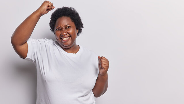 Fototapeta Photo of overjoyed dark skinned plump woman makes winner gesture raises arms and clenches fists feels like winner dressed in casual t shirt isolated over white background copy space for your advert