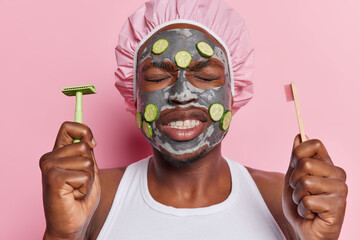 Horizontal shot of dark skinned man applies beauty mask with fresh cucumbers holds razor and...