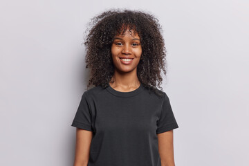 Waist up shot of pretty curly haired woman smiles happily dressed in casual black t shirt keeps...