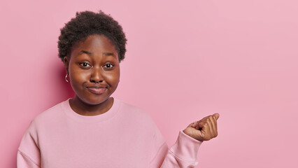 Horizontal shot of displeased overweight African woman with curly hair keeps hand raised up has chubby expression dressed in sweatshirt poses against pink background. Human facial expressions concept