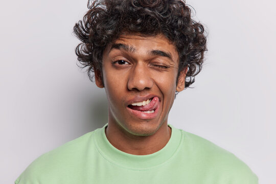Human facial expressions and fun concept. Funny curly haired Hindu man winks eye and sticks out tongue tries to make you laugh dressed in casual green t shirt isolated over white background.