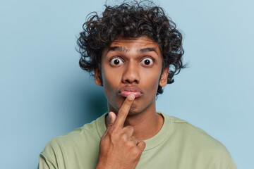 Portrait of Hindu man with curly hair keeps finger on lips stares bugged eyes with big surprisement...