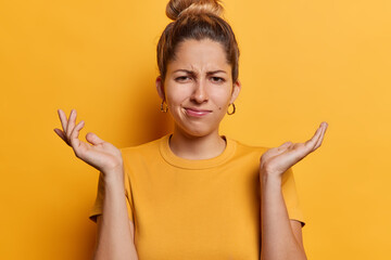 Clueless hesitant young woman with hair bun spreads palms feels unaware frowns face stands questioned dressed in casual clothes isolated over yellow background. Hm what to do in this situation - obrazy, fototapety, plakaty