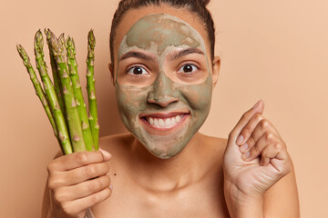 Photo of positive woman awaits for something clenches fist and holds bunch of green asparagus takes...