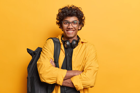 Horizontal Shot Of Handsome Curly Haired Hindu Man Dreessed In Casual Clothing Carries Black Rucksack On Shoulder Headphones Around Neck Isolated Over Vivid Yellow Background. Studying Concept