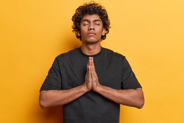 Photo of curly haired Hindu man keeps eyes closed prays and presses palms together believes in good...