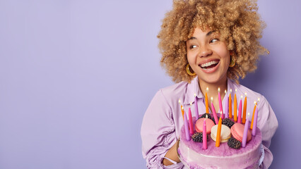 Studio shot of glad woman with curly bushy hair holds big tasty cake looks aside happily smiles...