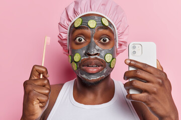 Photo of stunned dark skinned man applies beauty clay mask and cucumber slices holds toothbrush and smartphone undergoes daily hygiene procedures isolated over pink background. Wellness concept