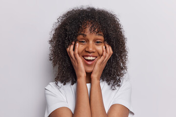 Portrait of cheerful curly haired woman keeps hands on cheeks laughs joyfully listens funny story...