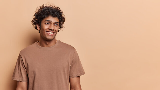 Waist up shot of handsome cheerful Hindu man with curly hair smiles positively concentrated aside reacalls funny moment in life wears casual t shirt isolated over brown background blank space for text