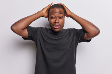 Waist up shot of confused shocked dark skinned man keeps hands behind head wears casual black t shirt purses lips has widely opened eyes isolated over white background reacts to something incredible