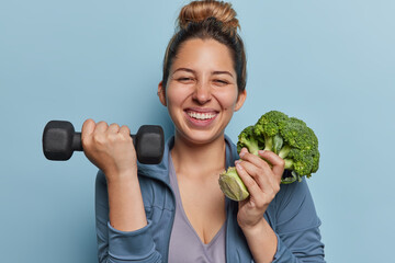Positive young active sportswoman leads healthy lifestyle lifts dumbbell and green broccoli keeps...