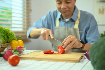 Shot of middle age man slicing ripe tomato on wooden board while cooking with organic food in...
